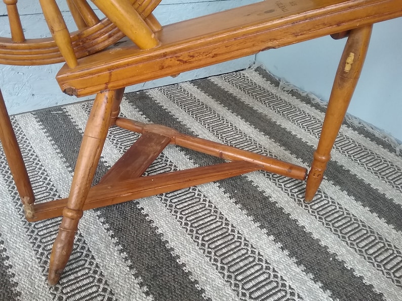 May include: A vintage wooden spinning wheel featuring a natural light brown finish with visible wood grain. The structure includes a large circular wheel, a horizontal frame, and three tapered legs connected by a triangular wooden base support.