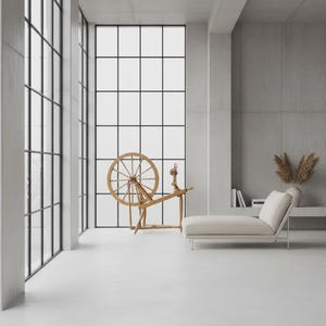 May include: A wooden spinning wheel stands in a room with large grid-patterned windows and concrete walls. Beside it, a light-colored chaise lounge sits on the floor. A vase with dried foliage and a few books are placed on a recessed shelf.