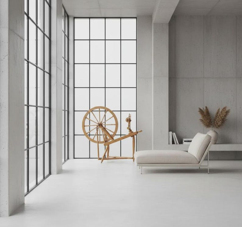 May include: A wooden spinning wheel stands in a room with large grid-patterned windows and concrete walls. Beside it, a light-colored chaise lounge sits on the floor. A vase with dried foliage and a few books are placed on a recessed shelf.