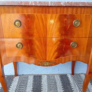 May include: A wooden antique chest of drawers with two drawers and a marble top. The chest has a warm, reddish-brown finish with brass hardware and decorative accents. The drawers have a symmetrical wood grain pattern.