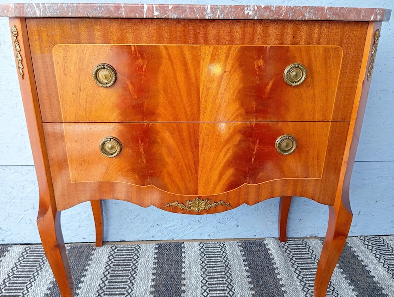 May include: A wooden antique chest of drawers with two drawers and a marble top. The chest has a warm, reddish-brown finish with brass hardware and decorative accents. The drawers have a symmetrical wood grain pattern.