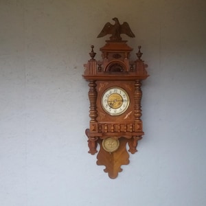 Könnte beinhalten: Eine antike Holzuhr mit einem Adler oben und einem Pendel. Das Zifferblatt der Uhr hat römische Ziffern und einen goldfarbenen Rand.