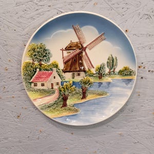 Puede incluir: Plato decorativo de cerámica con una escena de molino de viento holandés. El plato tiene un fondo de cielo azul con un molino de viento, una casa, árboles y una masa de agua. El molino tiene detalles marrones y rojos.
