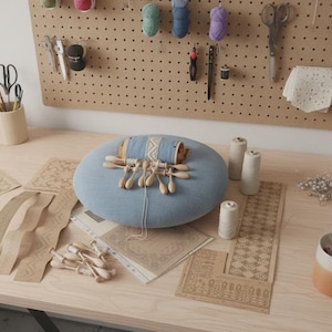 Può includere: Un cuscino circolare per merletti azzurro con fuselli in legno e filo, circondato da modelli in carta, rocchetti di filo bianco e una tazza bicolore. Il set include un pannello forato con vari strumenti e forniture artigianali.