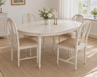 Puede incluir: Una mesa de comedor ovalada de color blanco roto con patas estriadas y cuatro sillas a juego con respaldos de listones verticales y asientos tapizados en tela estampada de color beige, azul y rojo. Un jarrón blanco con flores.
