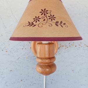 Op de afbeelding: Een houten wandlamp met een bruine lampenkap met een bloemenmotief. De lampenkap heeft een bordeauxrode rand.