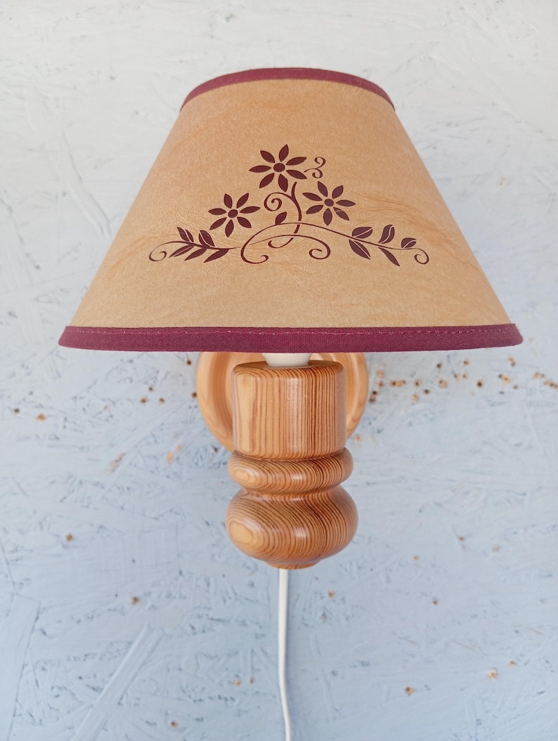 Op de afbeelding: Een houten wandlamp met een bruine lampenkap met een bloemenmotief. De lampenkap heeft een bordeauxrode rand.