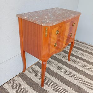 May include: A small, antique wooden chest of drawers with a marble top. The chest has two drawers with brass hardware and decorative accents. The wood has a warm, reddish-brown tone, and the legs are curved. The marble top has a pinkish hue.