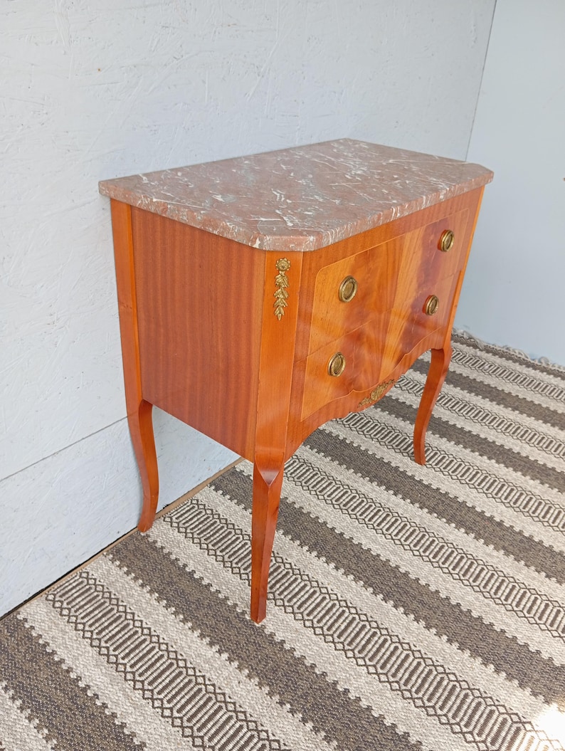 May include: A small, antique wooden chest of drawers with a marble top. The chest has two drawers with brass hardware and decorative accents. The wood has a warm, reddish-brown tone, and the legs are curved. The marble top has a pinkish hue.