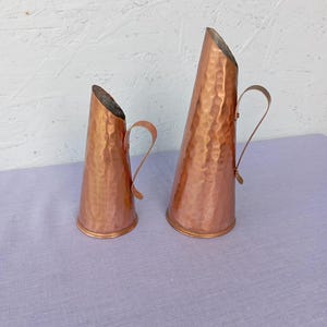Puede incluir: Dos jarras de cobre con texturas martilladas y asas curvas. Las jarras tienen forma cónica con la parte superior inclinada. El cobre tiene un tono marrón rojizo cálido. Una jarra es ligeramente más pequeña que la otra.