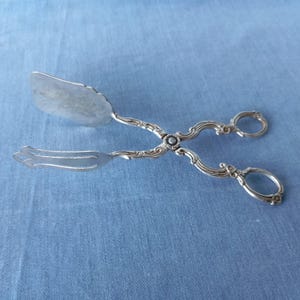 May include: Silver-plated serving tongs with an ornate design. The tongs have a flat, oval-shaped head and a decorative handle.