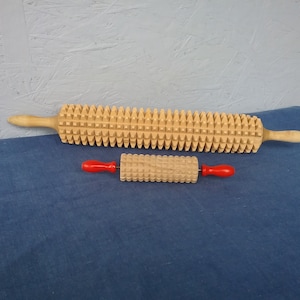 Puede incluir: Dos rodillos de masaje de madera. El rodillo más grande tiene un cuerpo largo y cilíndrico con una serie de protuberancias en forma de pirámide espaciadas uniformemente. El rodillo más pequeño tiene un diseño similar, con mangos rojos. Ambos son de madera clara.