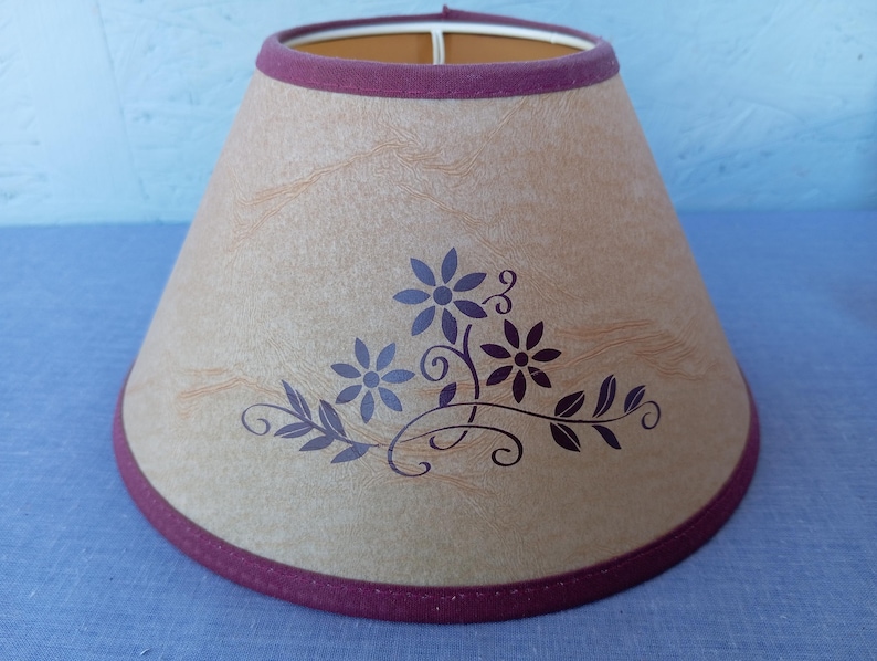 Op de afbeelding: Een beige lampenkap met een bordeauxrode rand en een bloemenmotief in donkerpaars. De bloemen zijn gestileerd en hebben vijf bloemblaadjes.