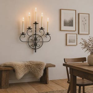 Puede incluir: Un aplique de pared de hierro forjado negro con cinco velas encendidas. El accesorio de metal tiene una columna central retorcida y adornos decorativos. El aplique está montado en una pared blanca, encima de un banco de madera con una piel de oveja.