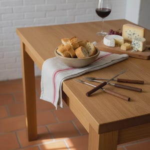 May include: A wooden table set for a meal, featuring a bowl of bread, cheese, grapes, and a glass of red wine. Several fondue forks with wooden handles are arranged on the table. A red and white striped cloth is also present.