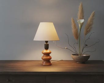 May include: A table lamp with a turned wooden base featuring a stacked, rounded design and a simple off-white conical fabric shade. The lamp emits a warm glow, illuminating the surface beneath it, and is positioned next to a small ceramic bowl.