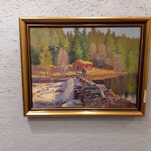 Könnte beinhalten: Ölgemälde in einem goldenen Rahmen, das eine rustikale Szene darstellt. Eine rote Hütte befindet sich in der Nähe eines Gewässers mit einer Holzbrücke. Der Hintergrund zeigt einen Wald mit grünen und gelben Bäumen unter blauem Himmel. Das Gemälde ist auf einer strukturierten, hellgrauen Oberfläche montiert.