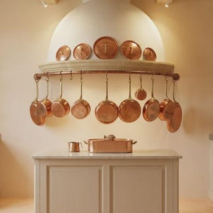 Op de afbeelding: Een kookeiland met een rechthoekige koperen braadpan en een kleine koperen beker. Daarboven hangt aan een gebogen koperen rek diverse koperen pannen, terwijl vijf koperen deksels op de rand van een grote witte afzuigkap zijn geplaatst.