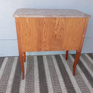 May include: A small, antique wooden cabinet with a light-colored marble top. The cabinet has a warm, reddish-brown wood grain finish and four curved legs. The cabinet is approximately 30 inches tall, 24 inches wide, and 18 inches deep.