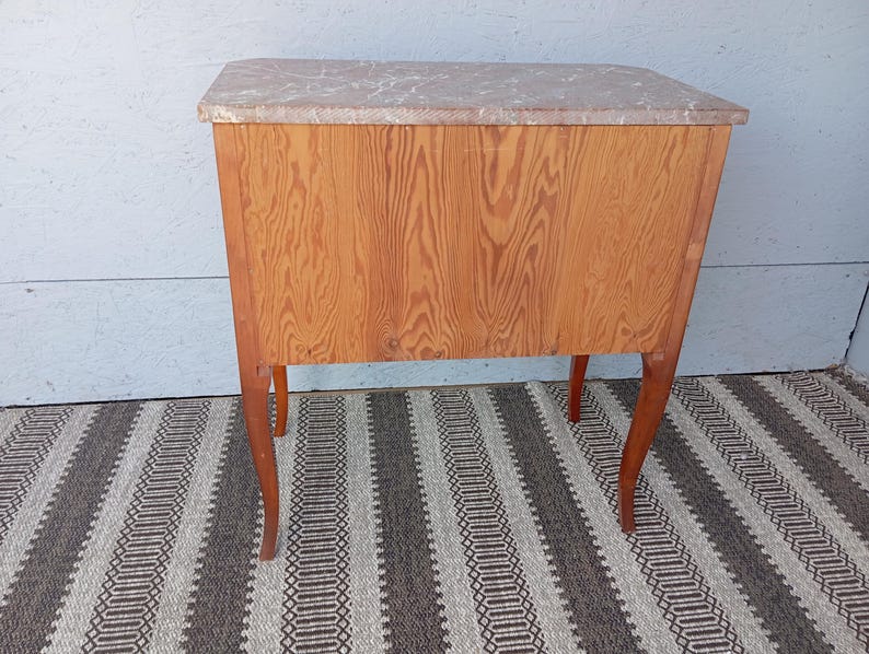 May include: A small, antique wooden cabinet with a light-colored marble top. The cabinet has a warm, reddish-brown wood grain finish and four curved legs. The cabinet is approximately 30 inches tall, 24 inches wide, and 18 inches deep.