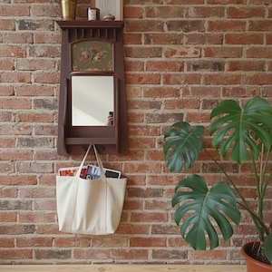 Puede incluir: Un espejo de pared marrón oscuro con un tapiz floral encima y una estantería. Una bolsa de mano blanca cuelga debajo del espejo, llena de revistas. El fondo es una pared de ladrillo rojo, con una planta en maceta a la derecha.