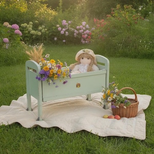 Può includere: Una culla per bambole in legno verde chiaro contenente una bambola di peluche con capelli di lana biondi e una cuffietta color crema. La culla è decorata con una piccola decalcomania floreale e contiene un bouquet di fiori di campo. Un cesto intrecciato.
