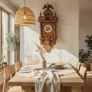 May include: A wooden wall clock with an eagle carving at the top, featuring a round face with Roman numerals. The clock is mounted on a wall above a light-colored wooden dining table set with a linen runner, a ceramic vase, and stacked plates.