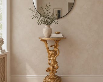 May include: A round gold pedestal table featuring a sculpted cherub figure as the central support base. The table has a circular top and rests on a decorative, ornate base with carved details. A round black-framed mirror hangs on the wall above.