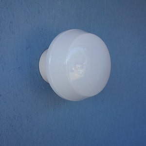 May include: A white, round, wall-mounted light fixture with a smooth, glossy finish. The fixture has a circular base and a larger, spherical shade. The background is a textured, blue surface.