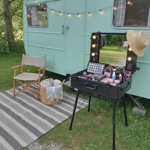 Può includere: Una postazione trucco portatile nera con specchio integrato e luci a LED, poggiata su quattro gambe regolabili. La valigetta è aperta e mostra varie palette di cosmetici, pennelli e flaconi. Un cappello di paglia è appeso alla cornice dello specchio.