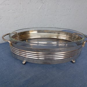May include: A silver serving dish with a glass insert and two handles. The dish has a decorative, ribbed design and sits on four small feet.