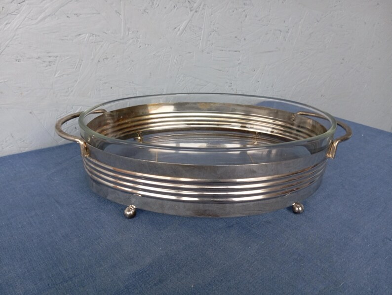 May include: A silver serving dish with a glass insert and two handles. The dish has a decorative, ribbed design and sits on four small feet.