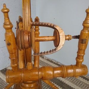 May include: A light brown wooden spinning wheel featuring a large vertical wheel, a horizontal flyer assembly with a U-shaped flyer arm, and two upright turned wooden posts. The mechanism includes a central bobbin and a series of metal hooks.