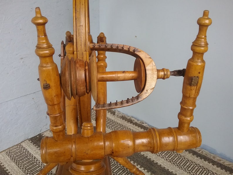 May include: A light brown wooden spinning wheel featuring a large vertical wheel, a horizontal flyer assembly with a U-shaped flyer arm, and two upright turned wooden posts. The mechanism includes a central bobbin and a series of metal hooks.