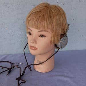 Puede incluir: Auriculares plateados vintage con un cable marrón, colocados sobre una cabeza de maniquí con pelo rubio corto. Los auriculares tienen un diseño circular y están conectados a un enchufe. El maniquí está sobre una superficie azul.