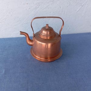 Könnte beinhalten: Eine Kupfer-Teekanne mit Deckel und Henkel. Die Teekanne steht auf einer blauen Oberfläche.