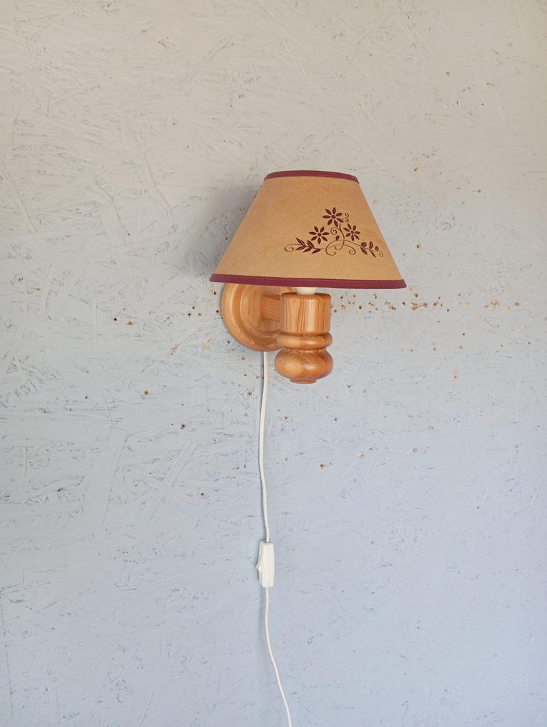 Op de afbeelding: Een houten wandlamp met een beige lampenkap met een bloemenmotief in donkerrood. De lampenkap heeft een donkerrode rand. De lamp heeft een wit snoer met een schakelaar.