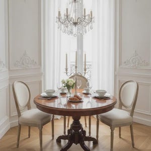 May include: A round wooden dining table with a pedestal base, surrounded by three upholstered chairs with light-coloured fabric. A silver candelabra, a glass vase with white flowers, and place settings with bowls and plates are on the table.