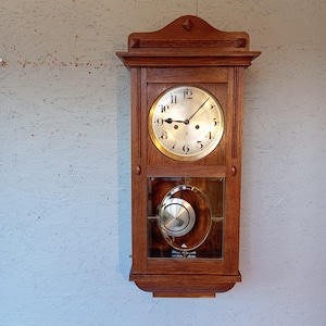 Puede incluir: Un reloj de pared de madera vintage con una esfera redonda dorada y manecillas negras. El reloj tiene un péndulo visible a través de una ventana de cristal. El marco de madera es de color marrón cálido y el reloj está colgado en una pared clara.