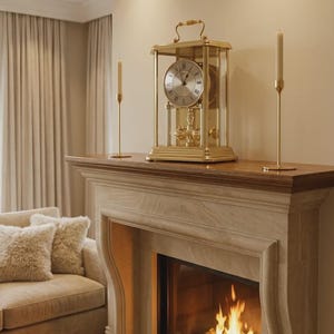 May include: A gold-toned mantel clock with a glass enclosure, a circular face featuring Roman numerals, and a visible pendulum mechanism, positioned between two slender gold candle holders on a wooden fireplace mantel shelf.