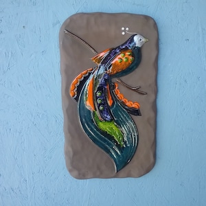 Puede incluir: Un adorno de pared de cerámica que presenta un pájaro estilizado con una cola larga y fluida. El pájaro está pintado en tonos de azul, verde, naranja y negro, y está posado en una rama. El fondo es una superficie marrón texturizada.