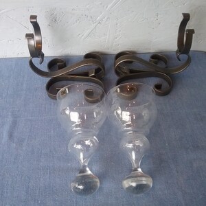 May include: A pair of black metal wall sconces with clear glass candle holders. The sconces have a decorative scroll design.