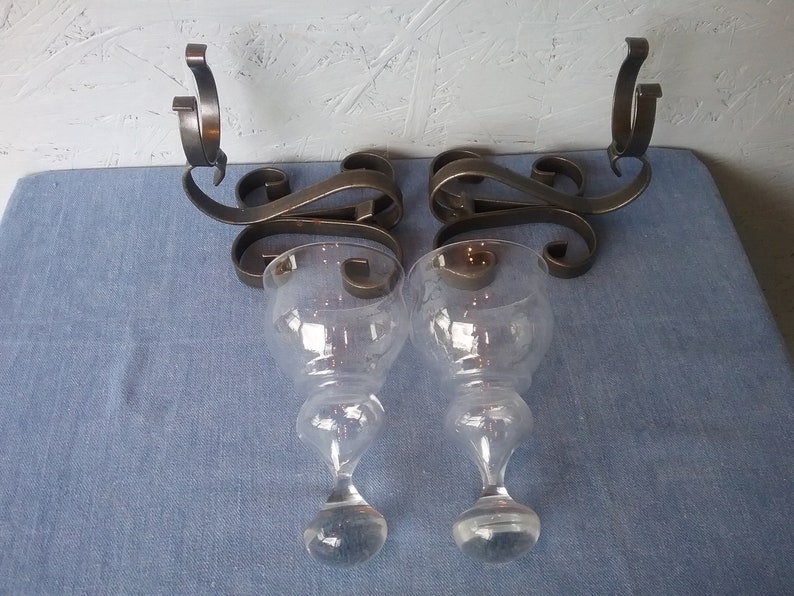 May include: A pair of black metal wall sconces with clear glass candle holders. The sconces have a decorative scroll design.