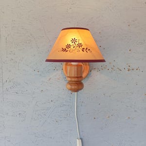 Op de afbeelding: Een houten wandlamp met een lichtbruine lampenkap van stof met een bloemenmotief. De lampenkap heeft een bordeauxrode rand. De wandlamp heeft een houten voet en een snoer met een schakelaar.
