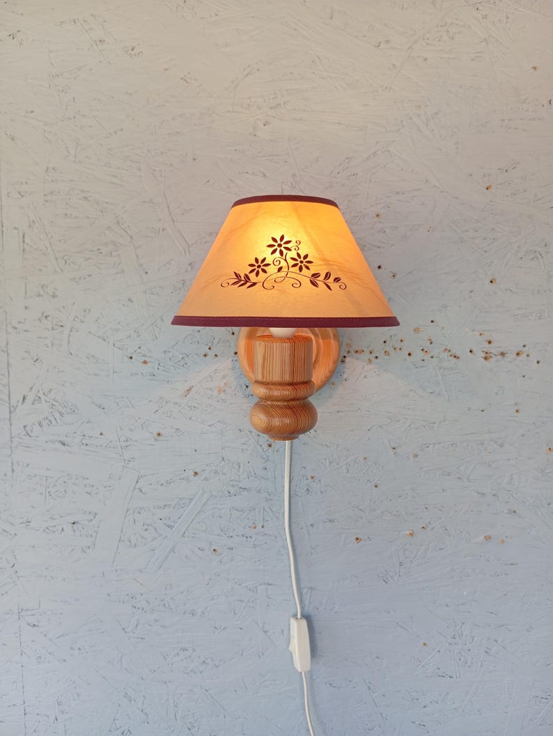 Op de afbeelding: Een houten wandlamp met een lichtbruine lampenkap van stof met een bloemenmotief. De lampenkap heeft een bordeauxrode rand. De wandlamp heeft een houten voet en een snoer met een schakelaar.
