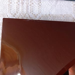 May include: A glossy, dark brown, triangular-shaped object with a smooth, reflective surface. The piece features a sharp, pointed corner and straight edges, showing subtle surface scratches and light reflections across its solid, dark-toned finish.
