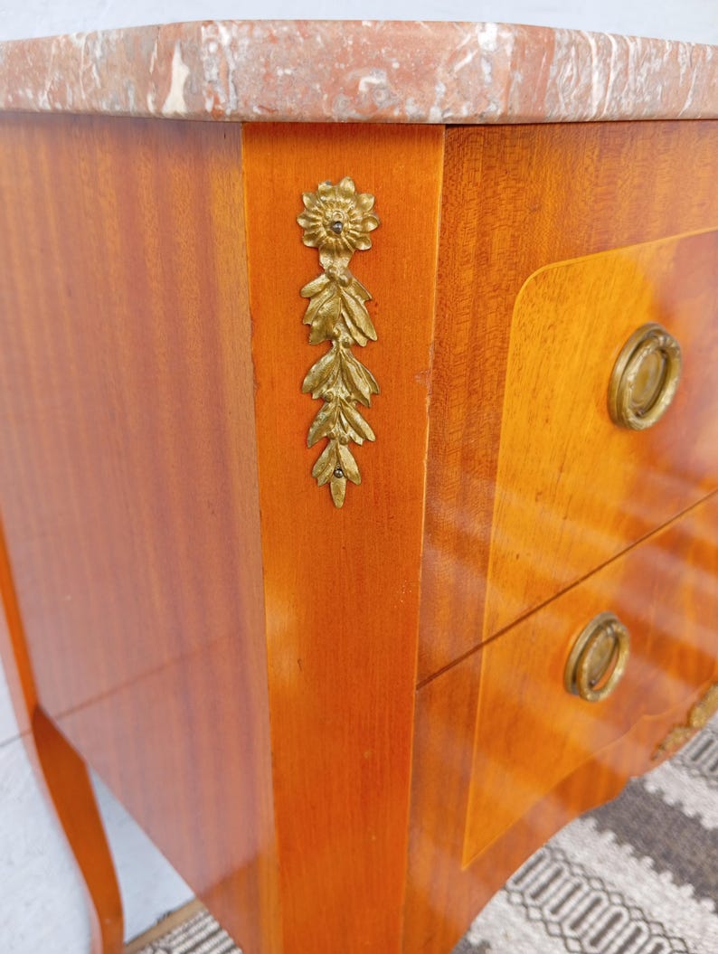 May include: A wooden nightstand with a marble top and brass hardware. The nightstand has two drawers and decorative brass floral accents. The wood has a warm, reddish-brown finish.