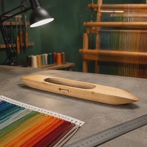 May include: A light brown wooden weaving shuttle on a grey surface, with a rainbow colour palette of threads in the foreground. A ruler and a weaving loom are also visible in the background. The shuttle is a light brown colour.