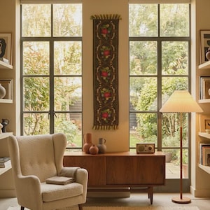 Puede incluir: Un sillón tapizado de color crema con respaldo capitoné, un aparador de madera, un tapiz vertical con bordados florales, una lámpara de pie con pantalla plisada y una alfombra texturizada frente a dos grandes ventanales.