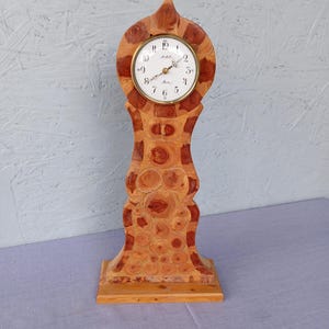 May include: A wooden mantel clock with a light brown finish and a circular clock face. The clock face has black numerals and gold-coloured hands. The clock is approximately 38 cm tall and has a unique, decorative design.